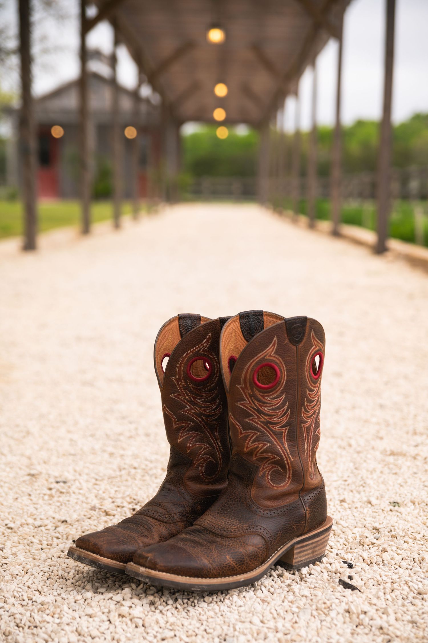 Rustic brown leather cowboy boots on a gravel path with covered walkway in background.