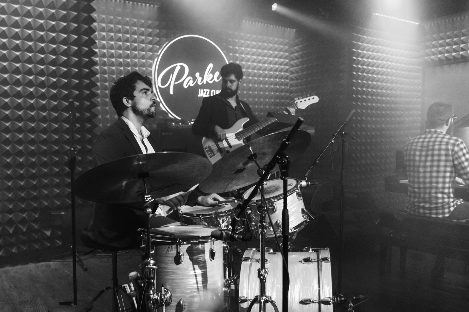 Musicians play drums and guitar in black and white at Parker Jazz Club.