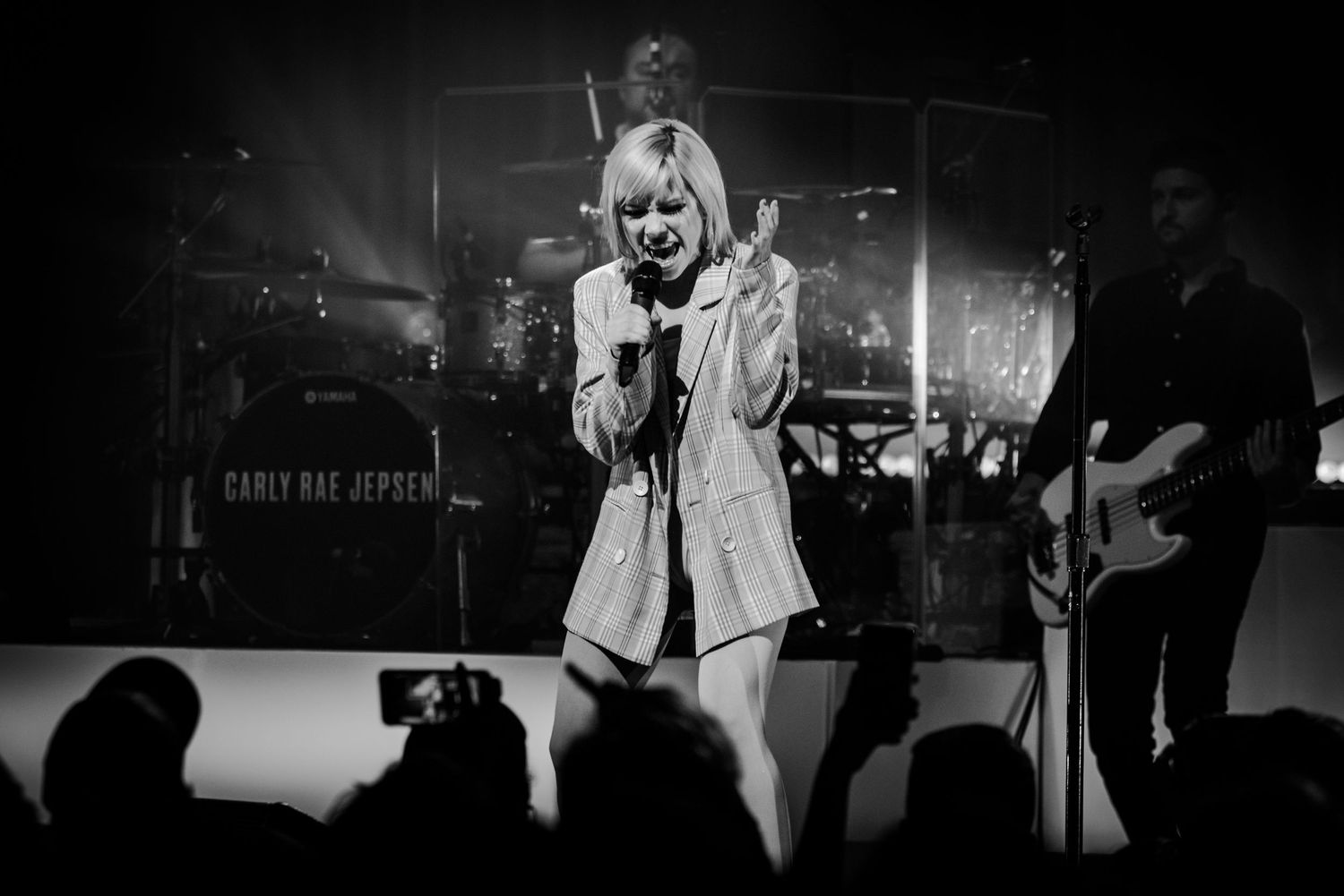 Concert performer in white outfit singing on stage with band members in dark silhouettes.