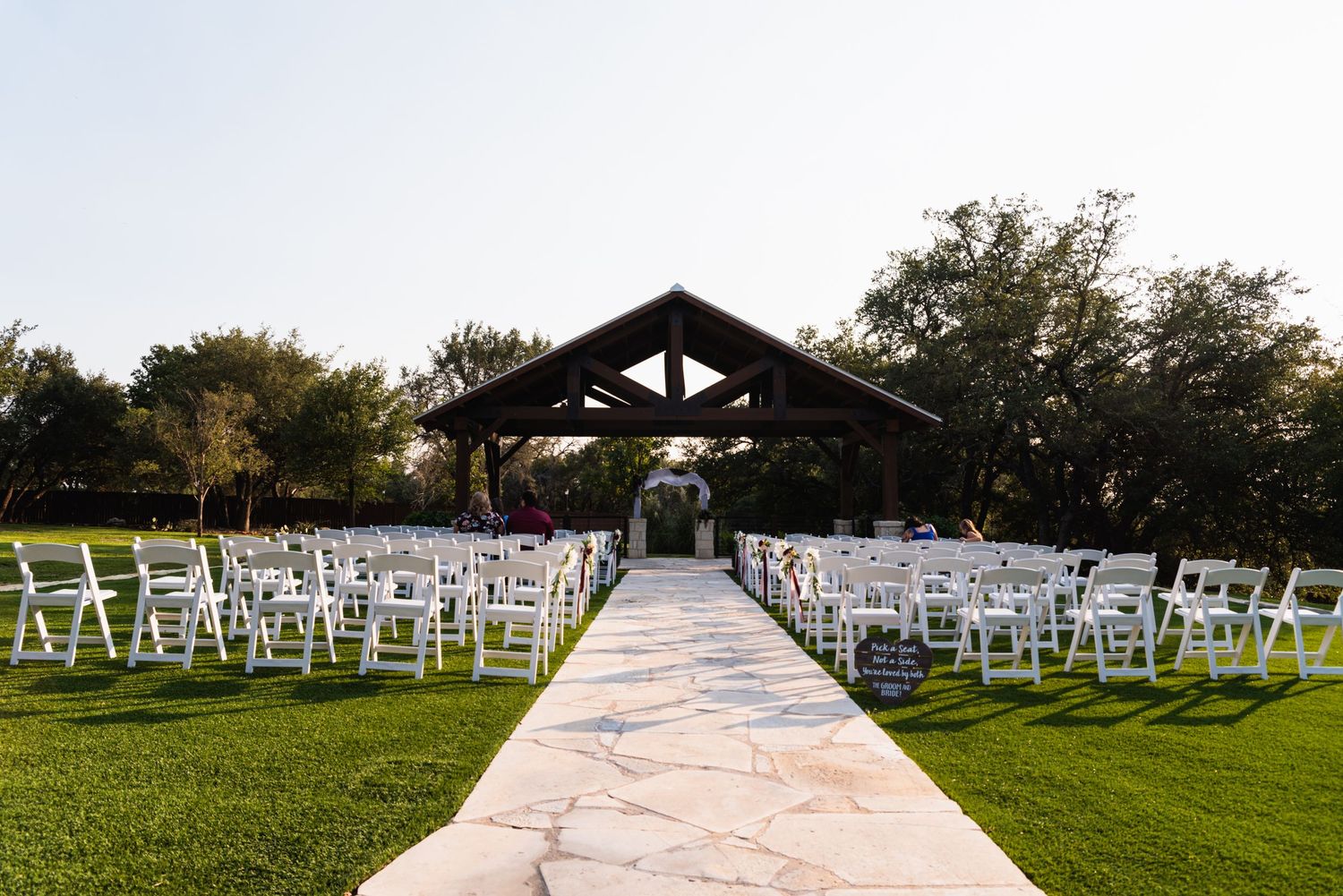 White chairs arranged for an outdoor wedding ceremony leading to a rustic wooden pavilion on manicured lawn.