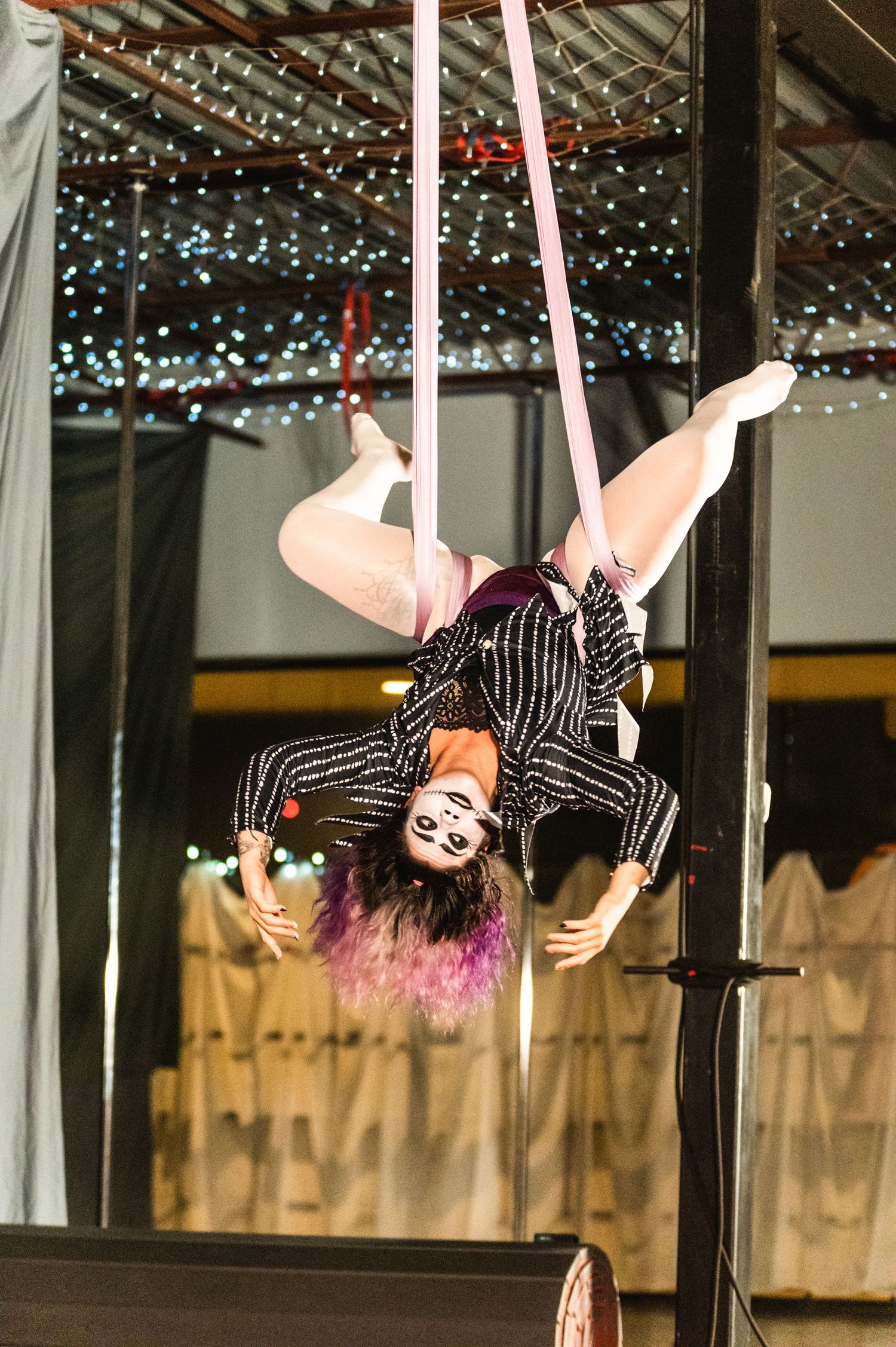 Aerial artist in striped outfit performs upside down on pink silks under twinkle lights.