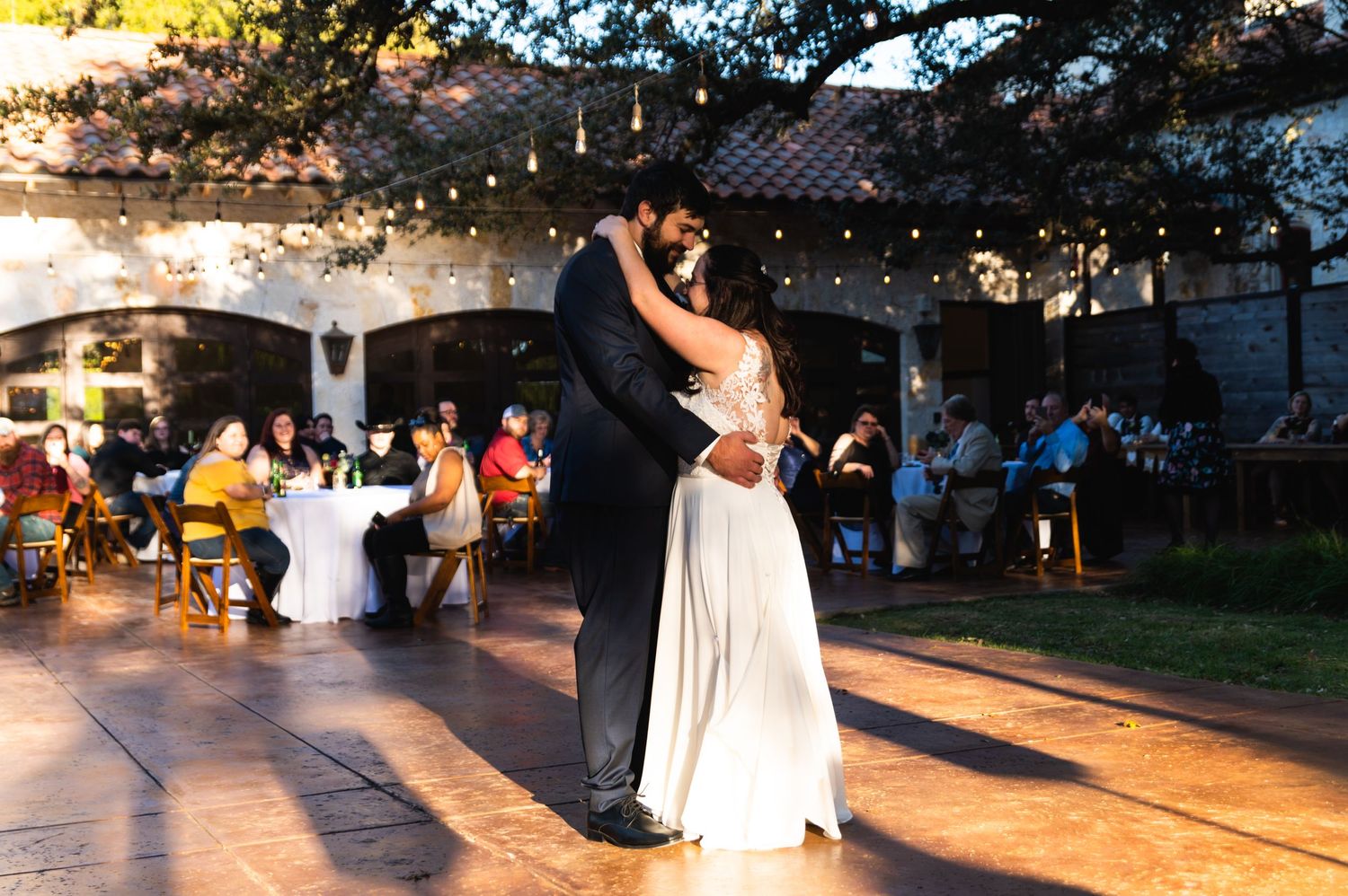 A couple dances intimately at their wedding reception as string lights twinkle overhead at dusk.