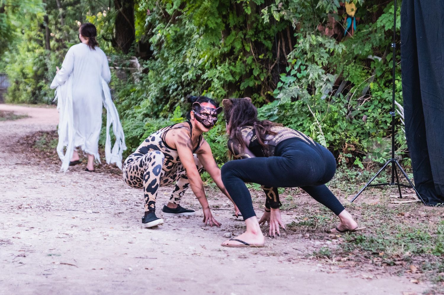 Performers in costume crawl on dirt path with figure in white in background.