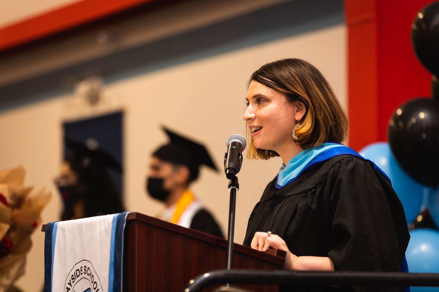 Speaker at graduation podium delivering address with blue stole and black academic regalia.