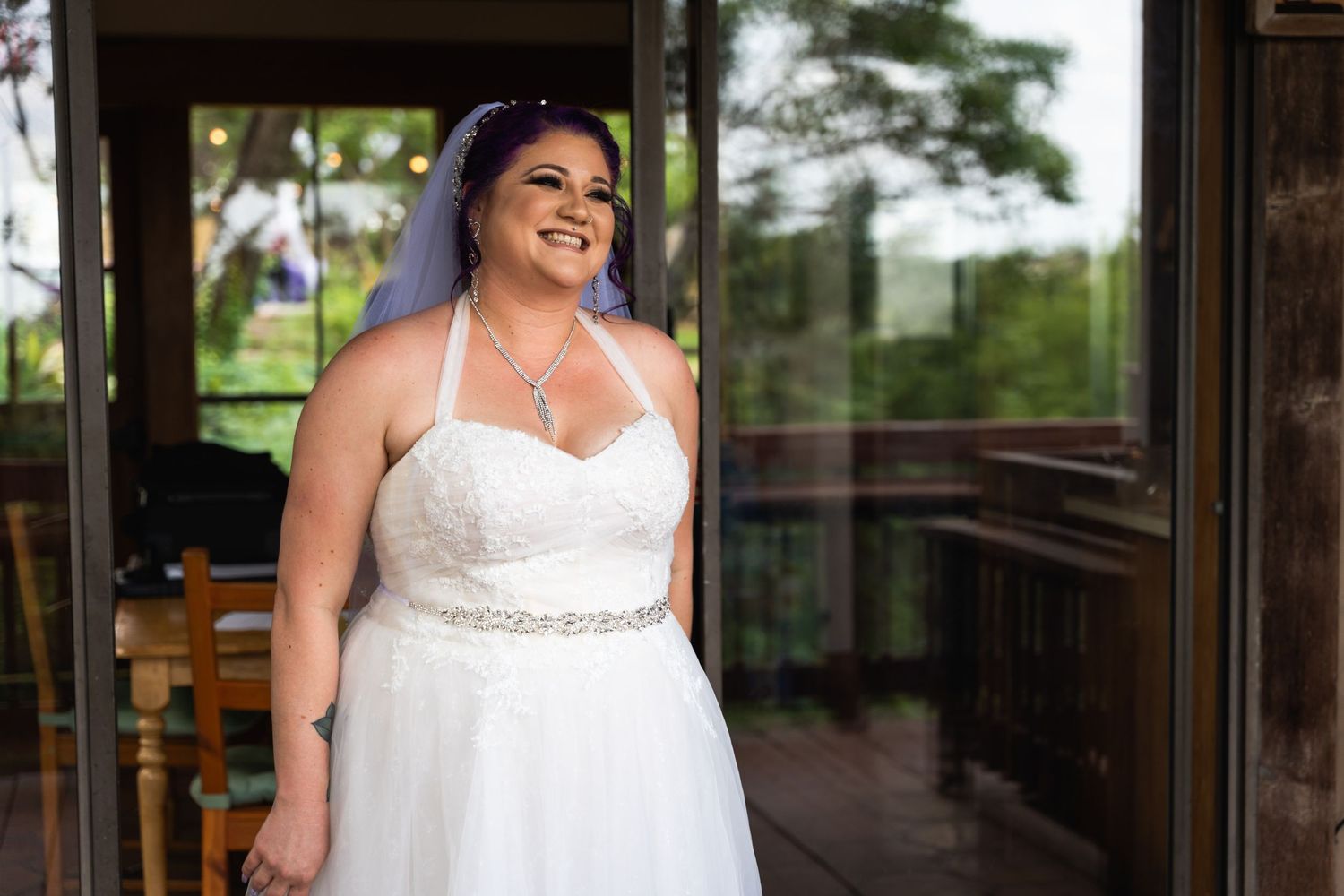 A bride in a strapless white wedding gown stands smiling in a rustic outdoor setting.