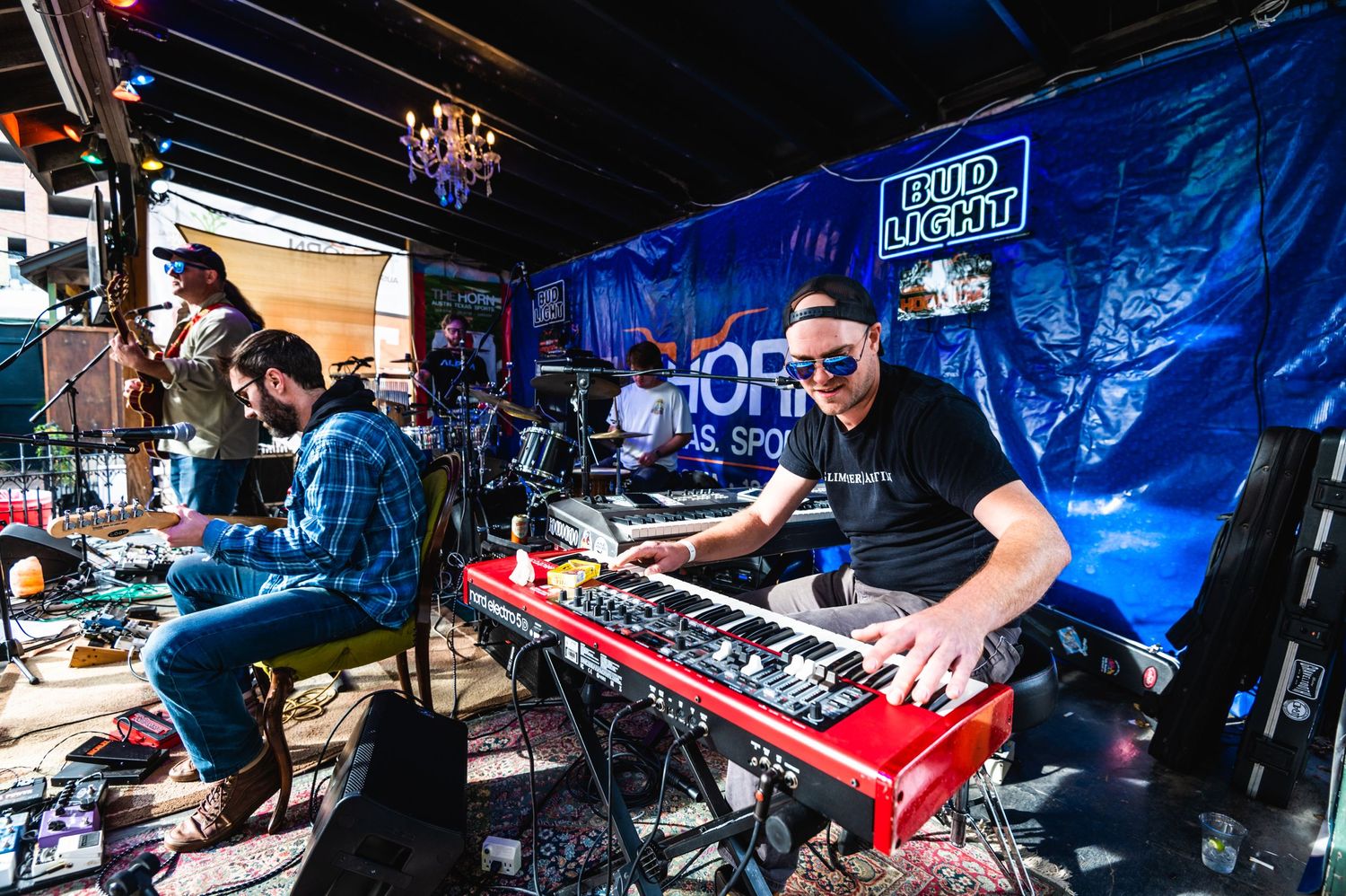 Musicians perform on stage with keyboard and instruments against blue backdrop at live music venue.
