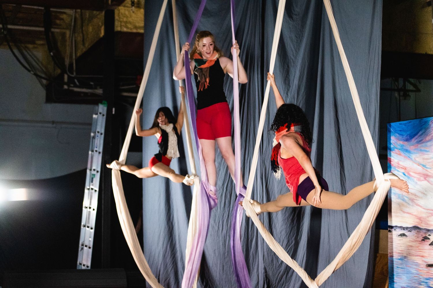 Three aerial artists perform synchronized poses on white and purple silks in a dimly lit studio.