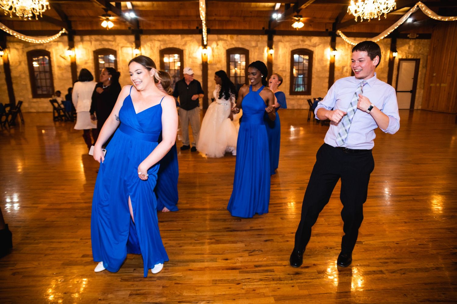 Guests in blue attire dance energetically on a wooden dance floor under crystal chandeliers.