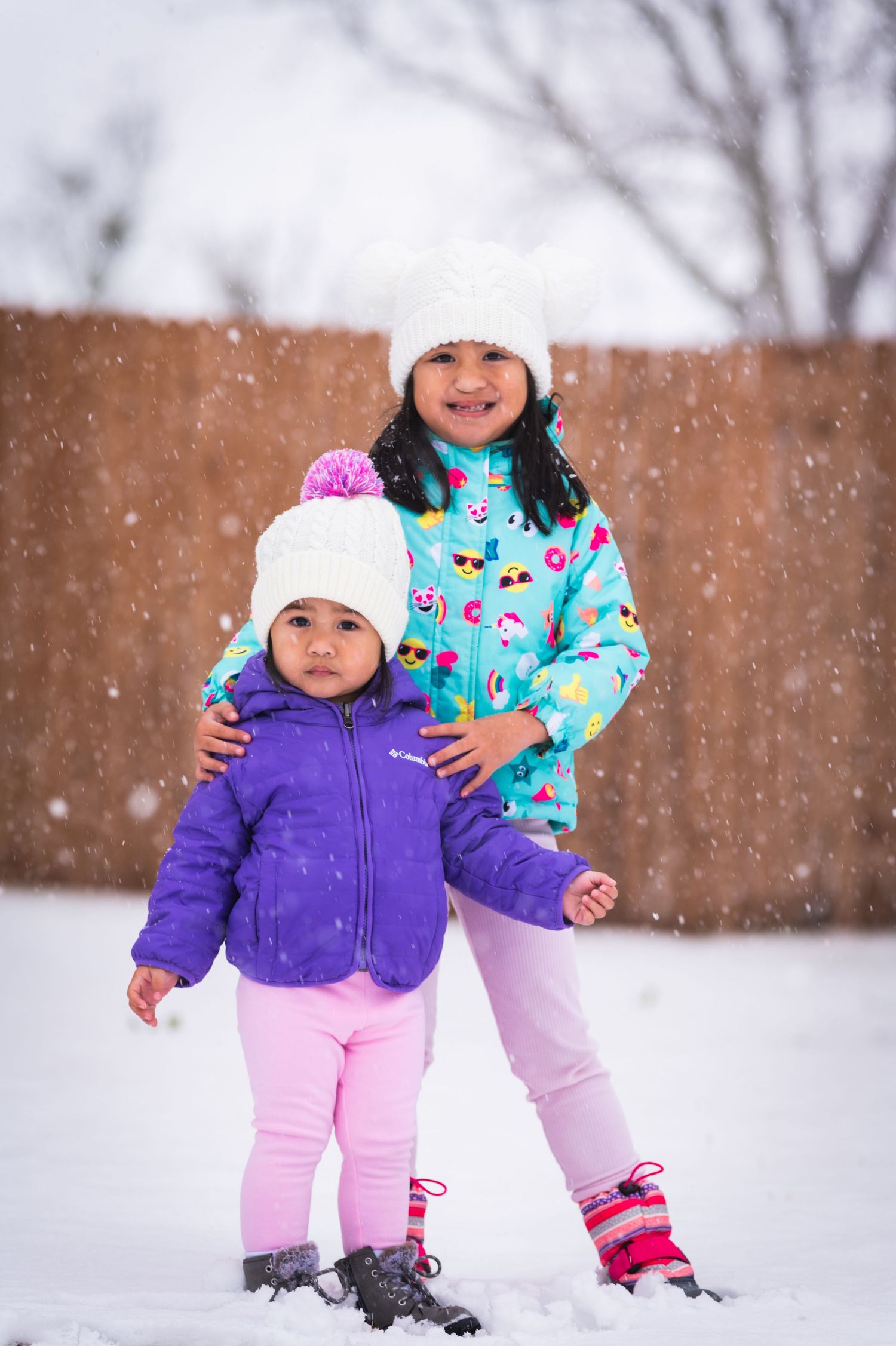 Two bundled up children pose together in the falling snow wearing colorful winter coats.