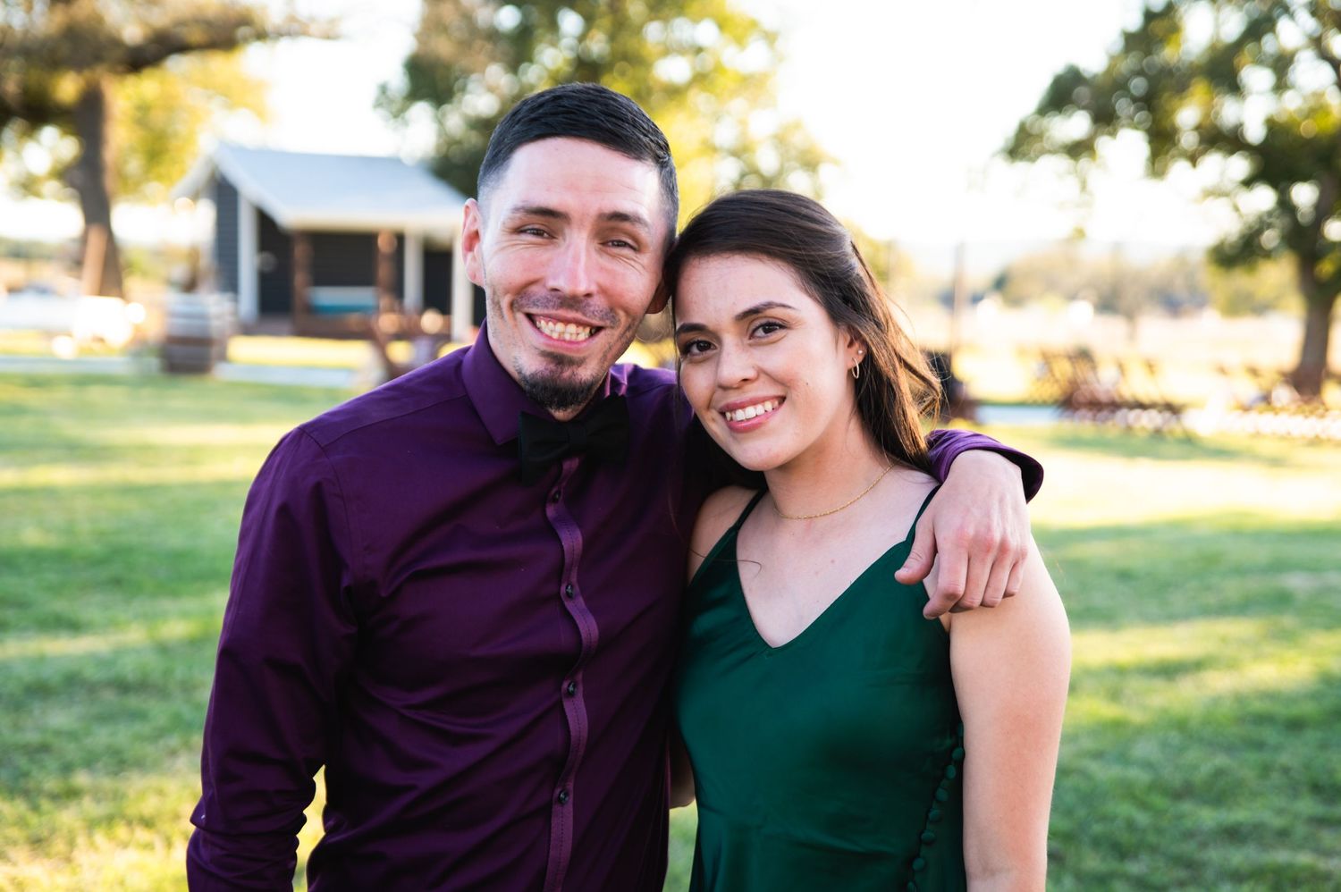 A couple poses together outdoors on a sunny day with green grass and buildings in background.
