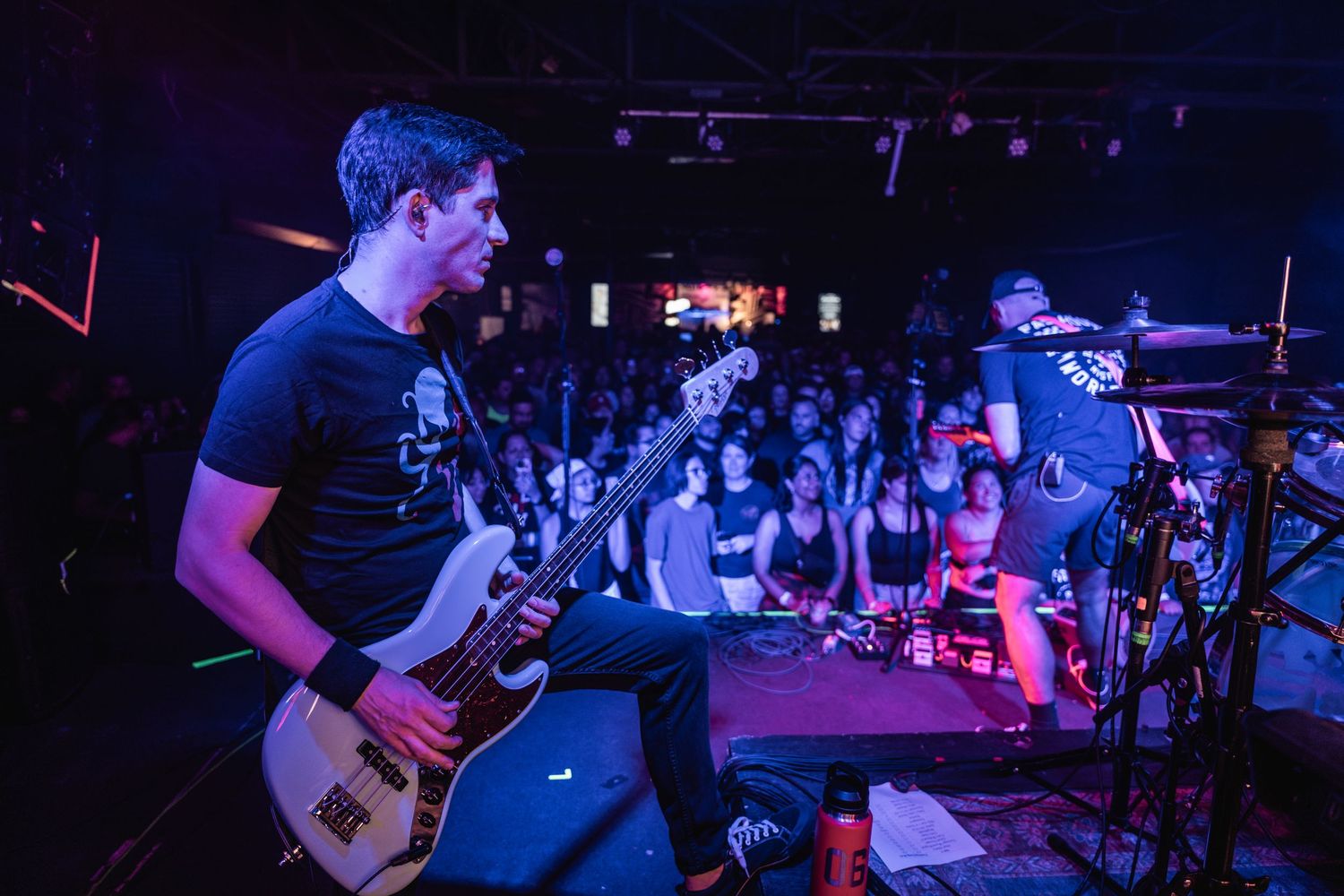 Band performs energetically on stage at a dark concert venue with purple lighting and crowd visible in background.