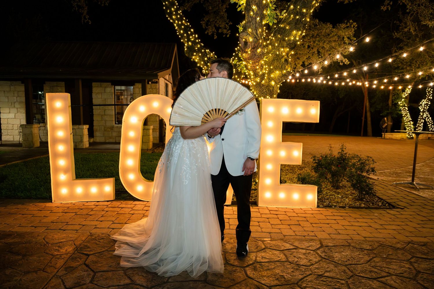 A couple embracing in front of illuminated LOVE marquee letters at night during their wedding celebration.