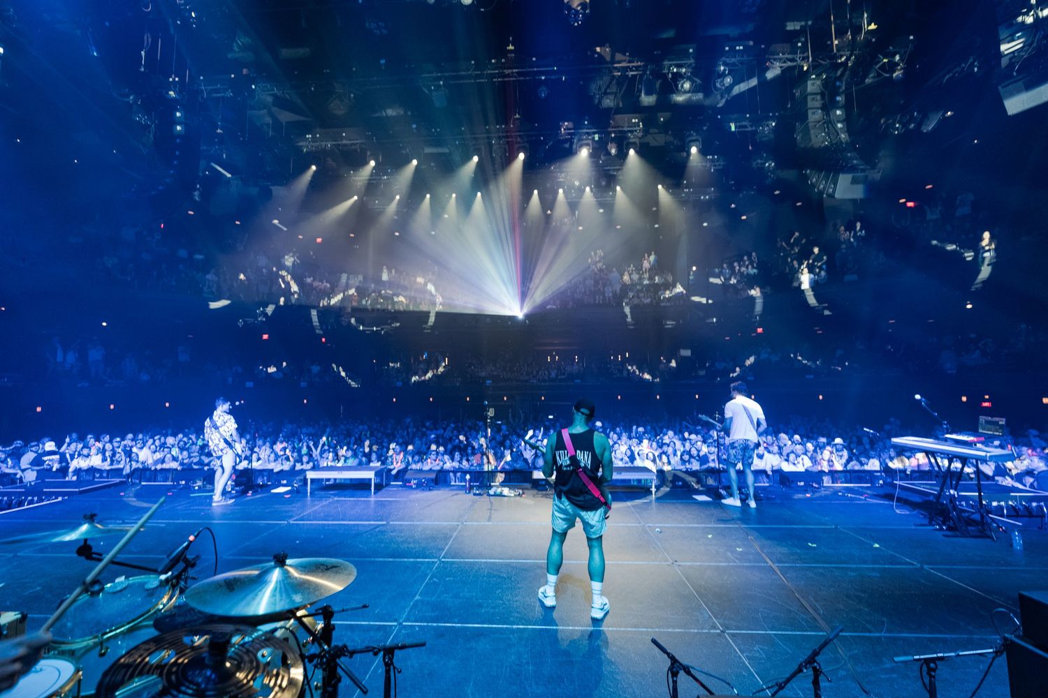 Rock band performs on a massive illuminated stage with dramatic blue lighting and laser beams before a packed arena crowd.