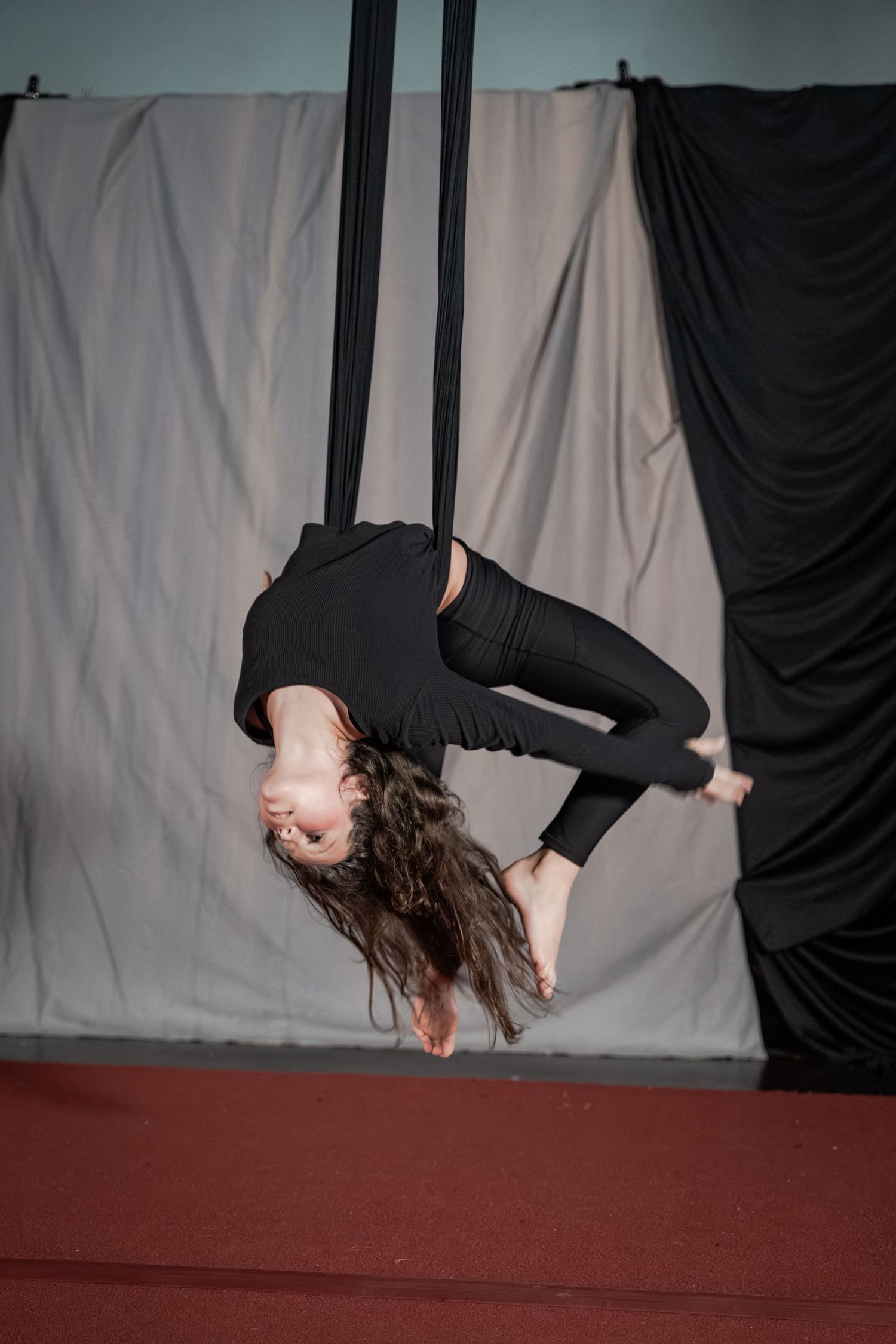 Aerial silk performer in black outfit hanging upside down from fabric ribbons in a studio setting.