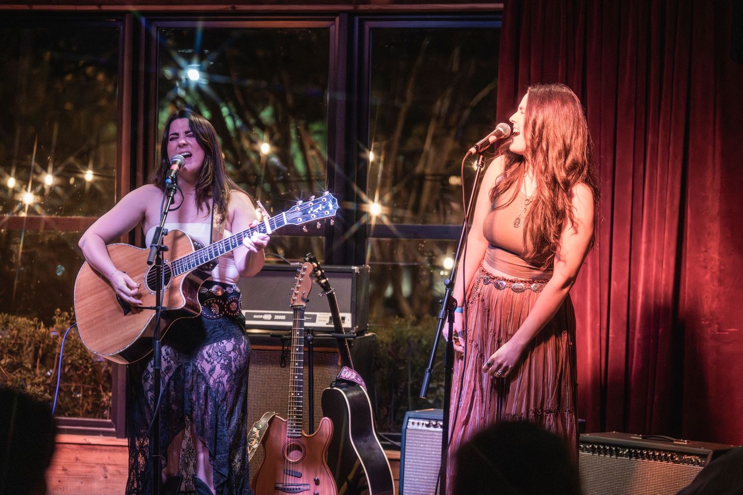 Musicians perform on stage with guitars and microphones at a live music venue with red curtain backdrop.