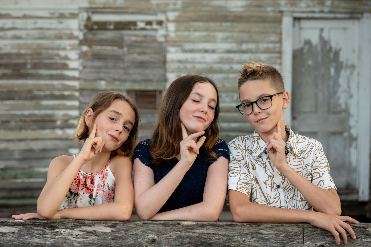 Three siblings posing playfully while leaning on a wooden surface outdoors.