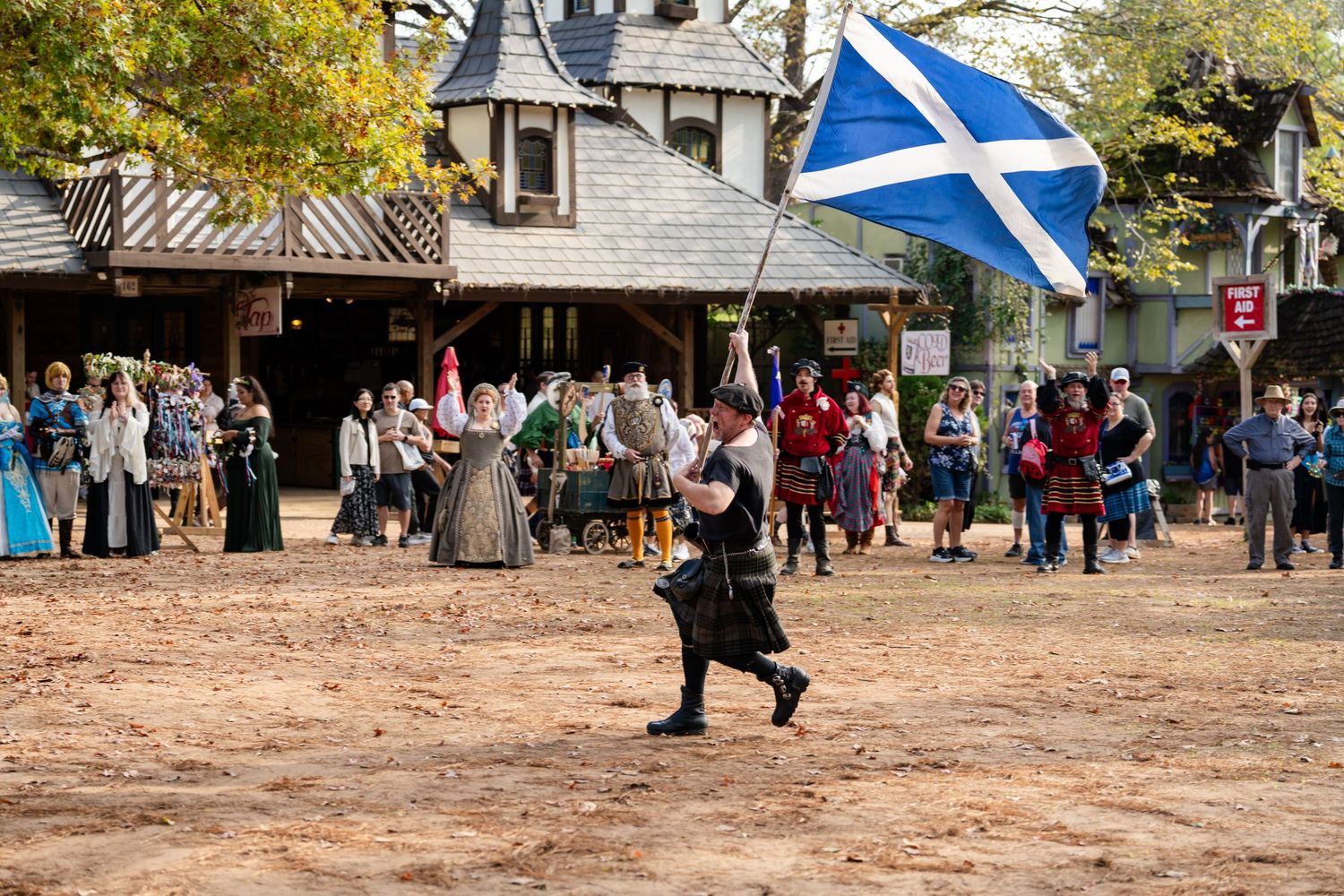 A person runs with a Scottish flag at a Renaissance festival while crowds gather around rustic buildings in the background.