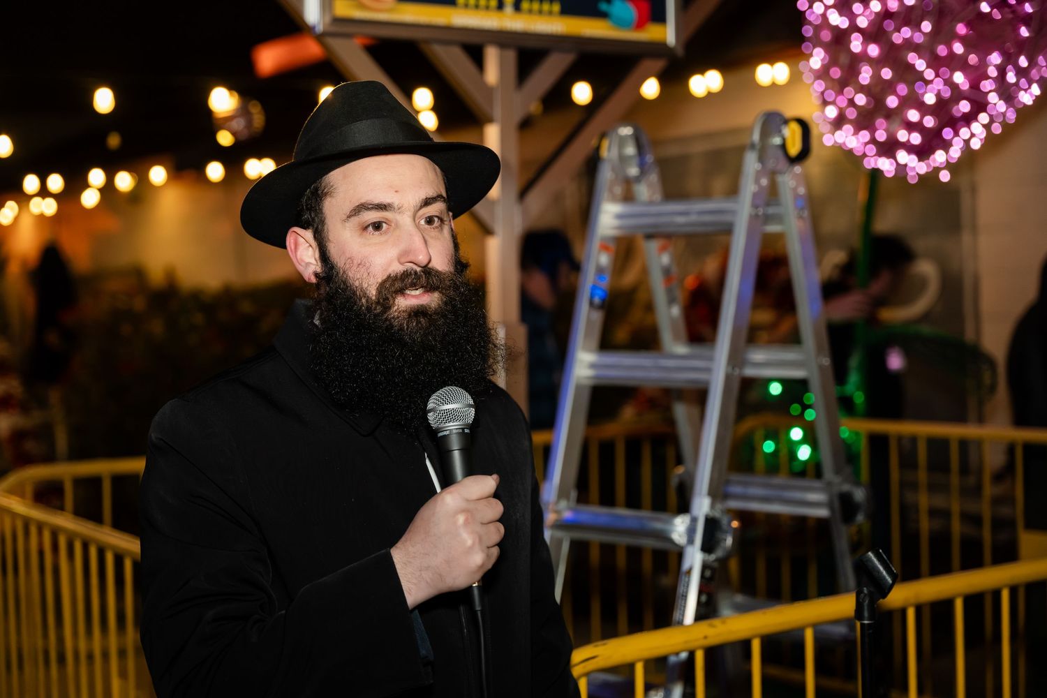A performer in a black hat holds a microphone while standing on stage with colorful lights in the background.