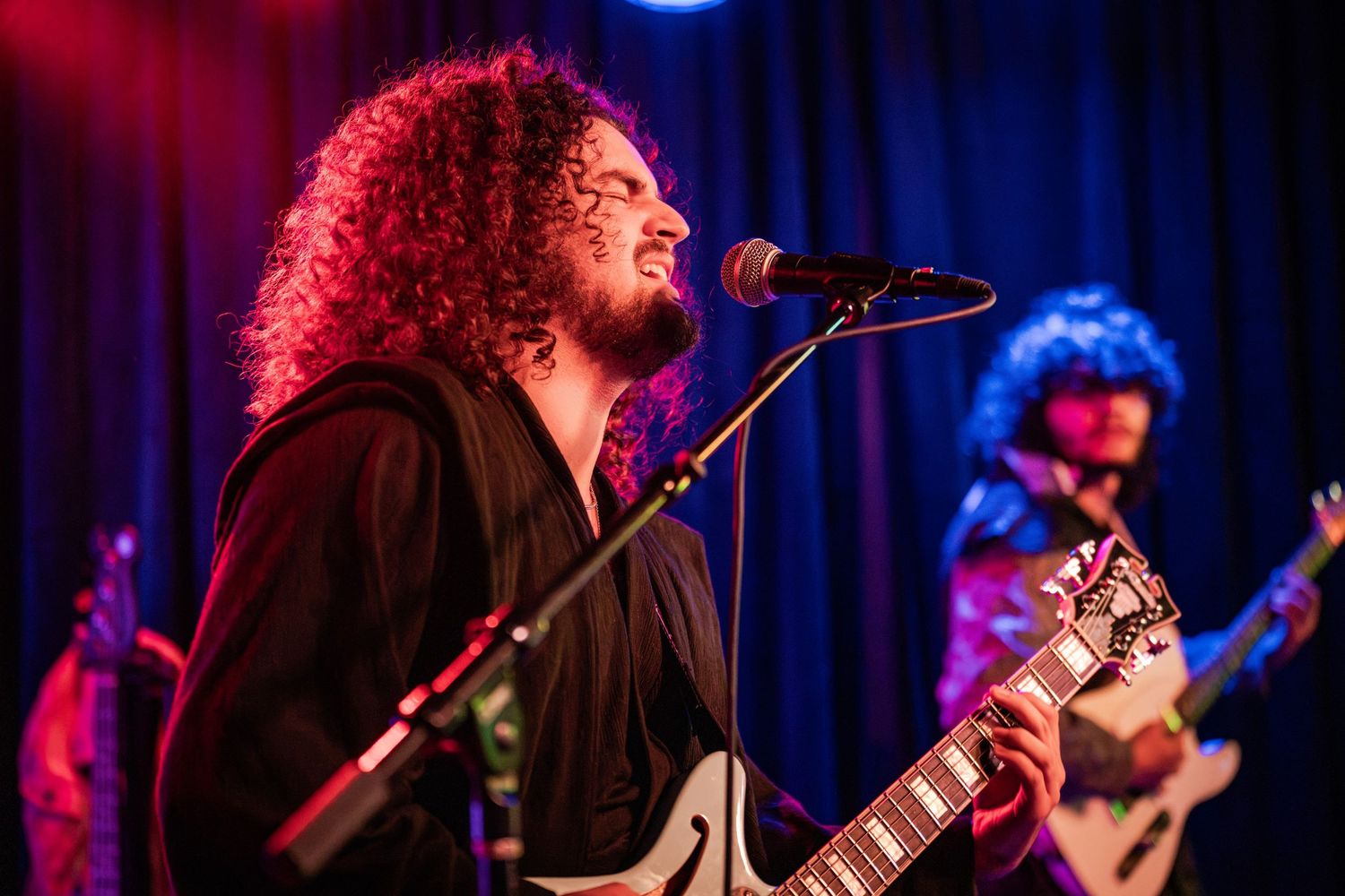Musicians perform on stage with electric guitars under dramatic blue and red lighting during a live concert performance.