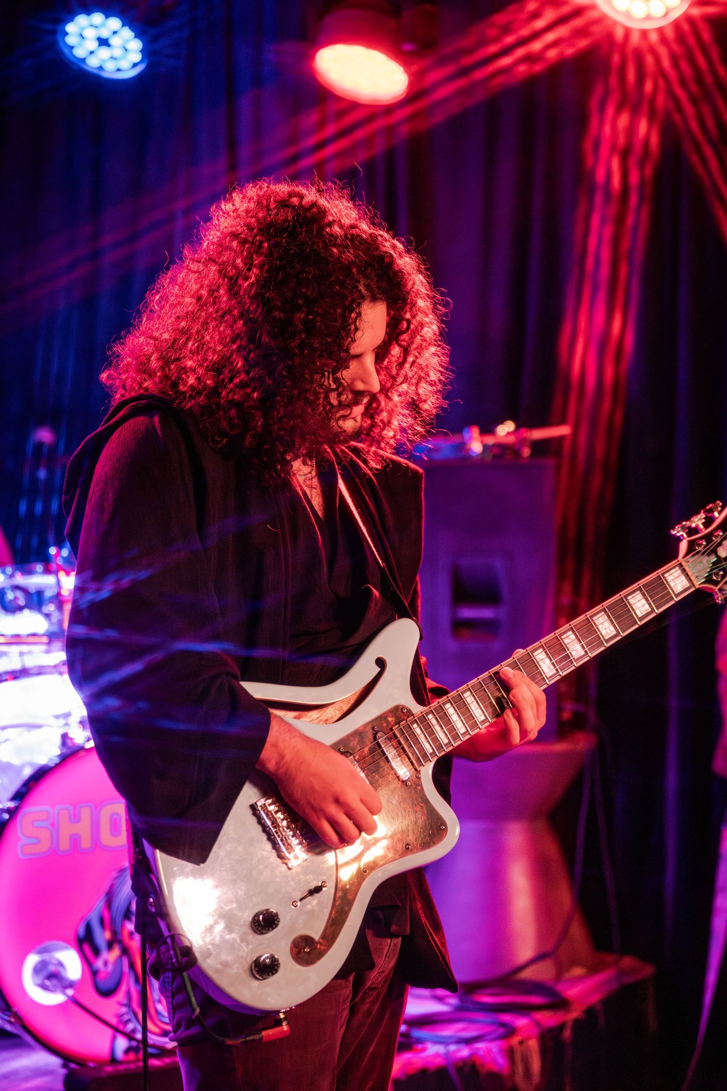 Guitarist with curly hair performs on stage under dramatic red and blue lighting while playing a white electric guitar.
