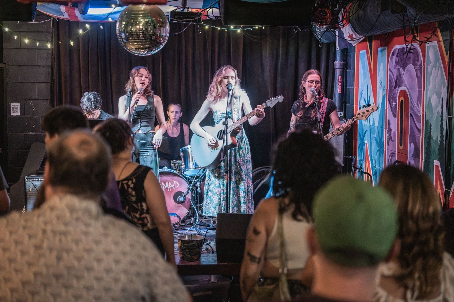 A crowd watches a live band perform on stage in a dimly lit venue with colorful lighting effects.