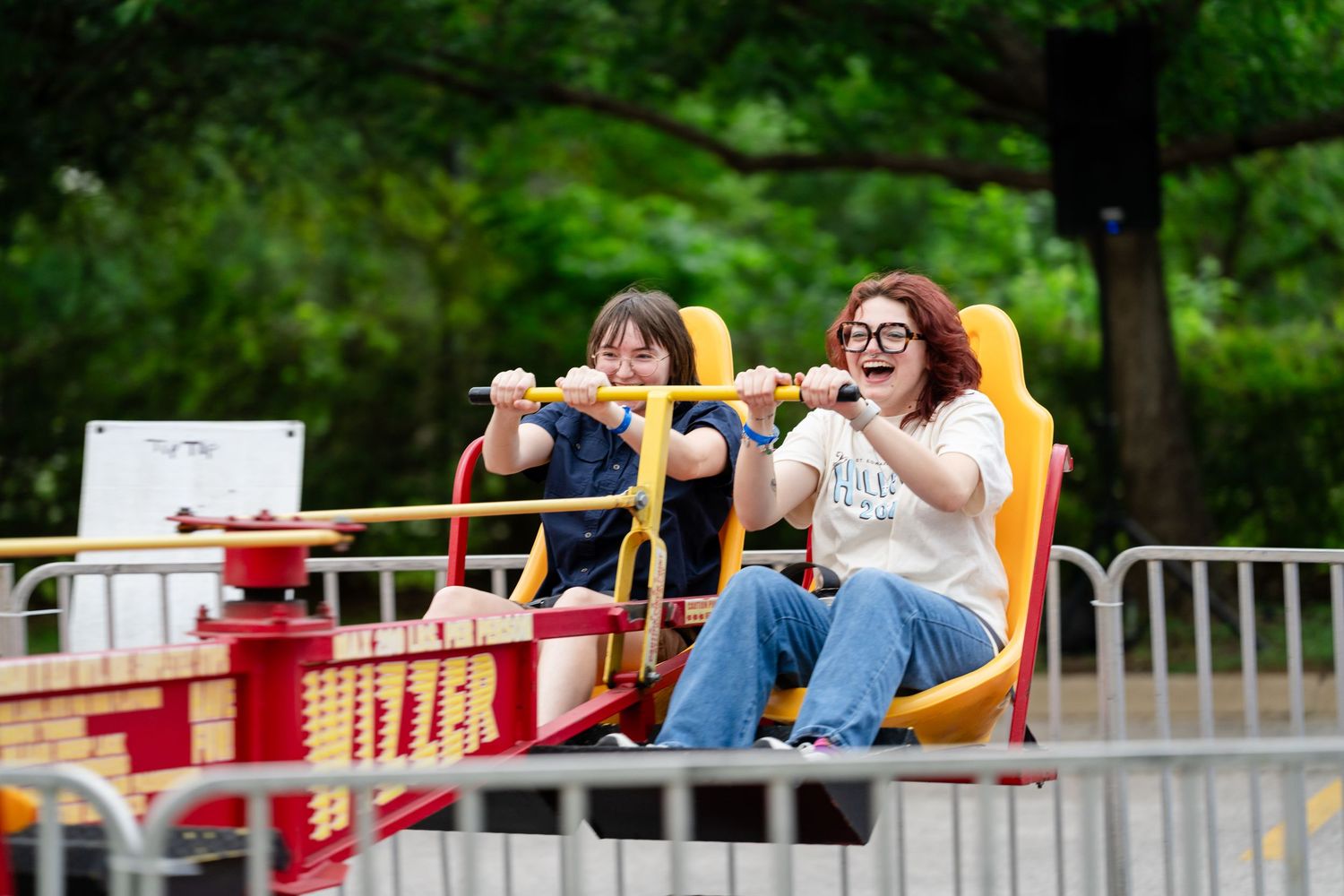 Two women laughing and enjoying a carnival ride at an outdoor amusement park surrounded by green trees.