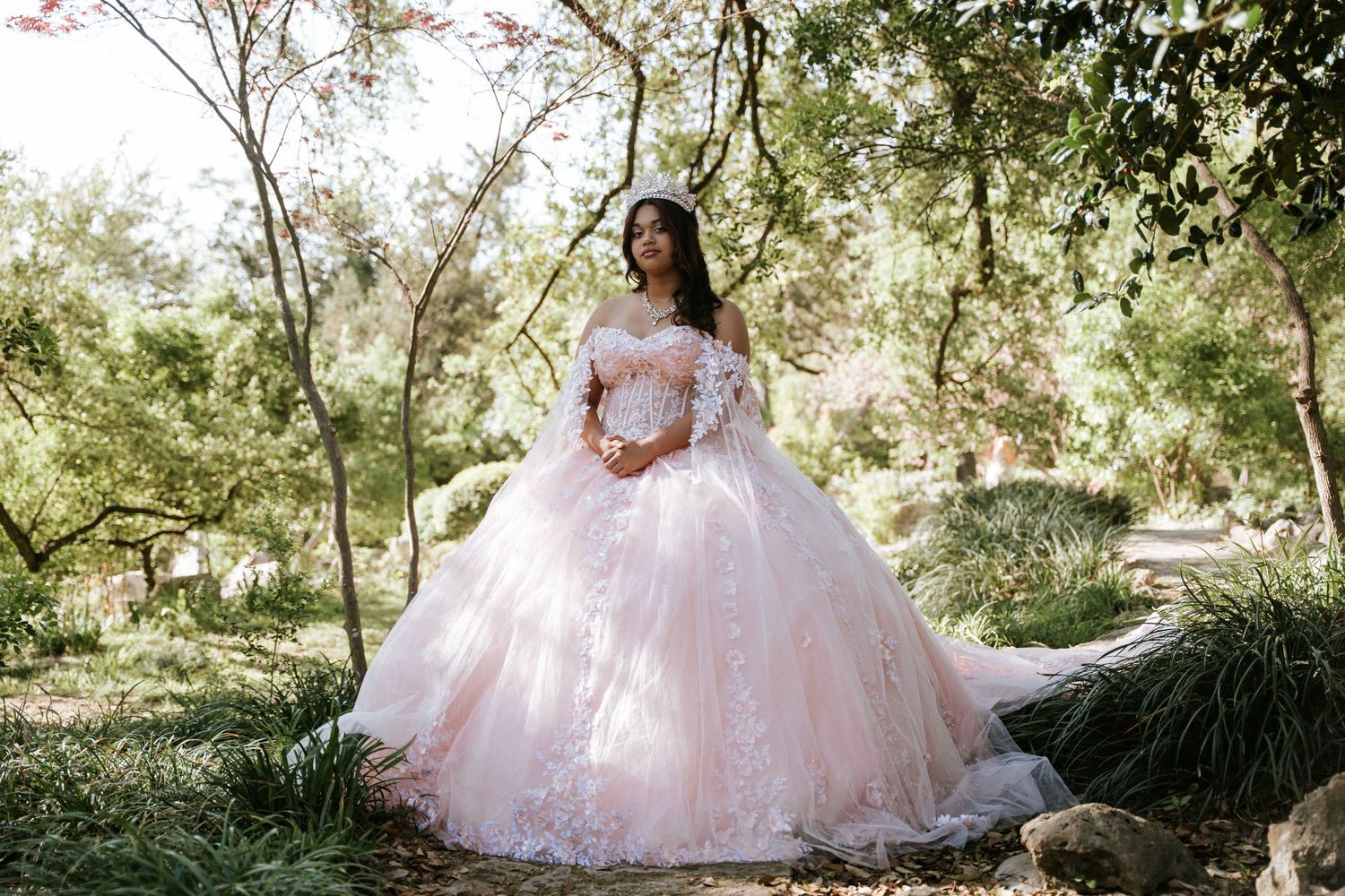A woman in a blush pink ball gown with lace details poses gracefully in a lush garden setting.