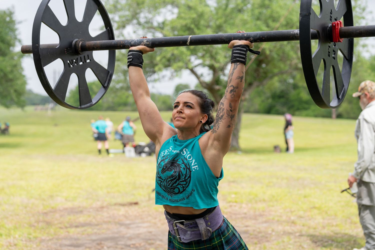Woman lifting barbell overhead outdoors at a strongman competition wearing a teal shirt and plaid skirt.