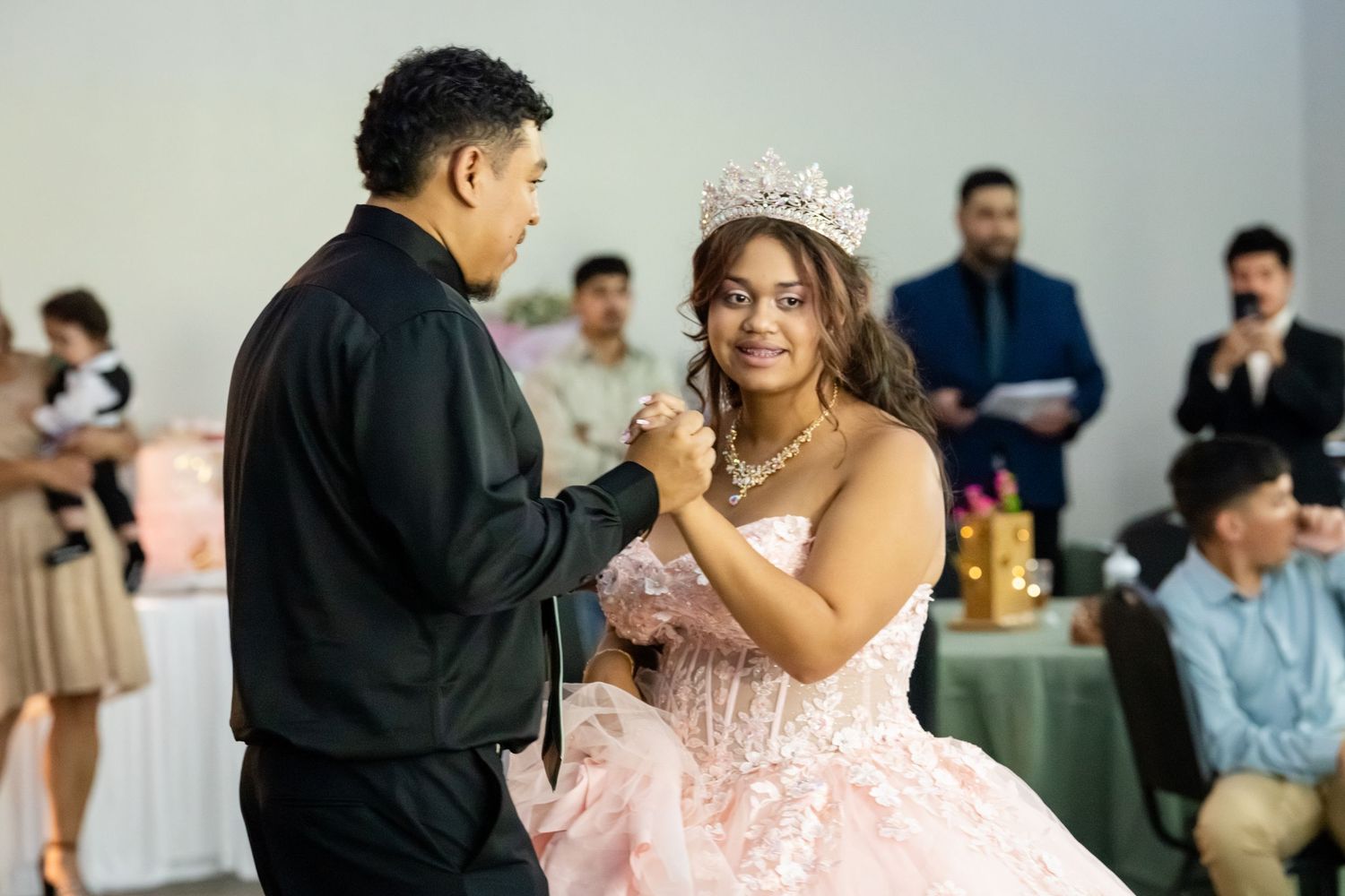 A young woman in a pink quinceañera gown and tiara shares a dance with a man in a black jacket at her celebration.