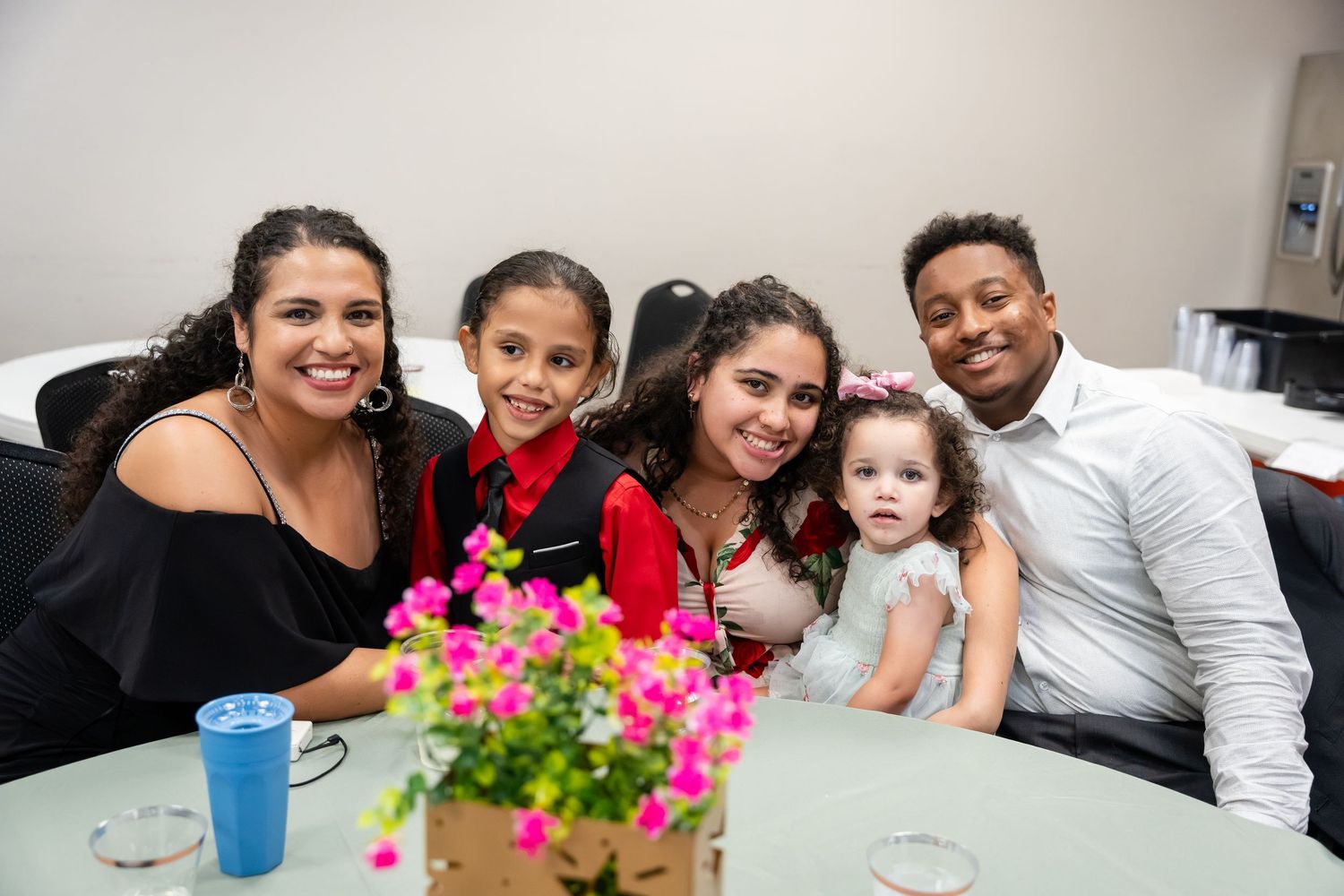 Happy family of five posing together at a table with pink flowers, smiling warmly in a bright indoor setting.