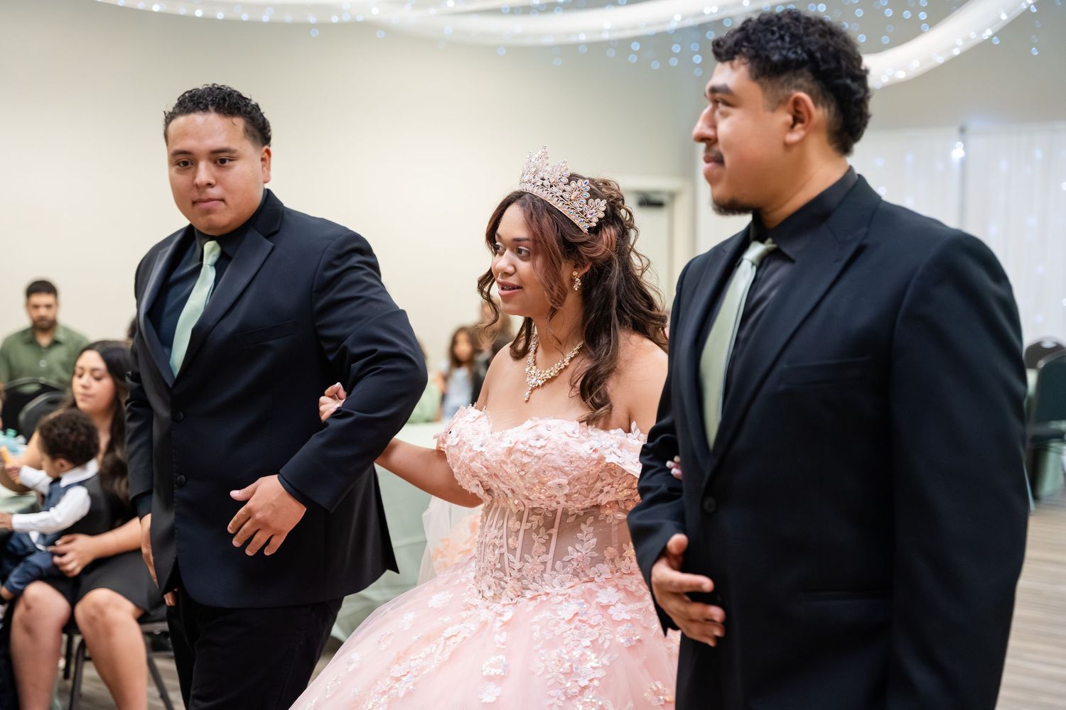 Young woman in pink quinceañera gown escorted by two men in black suits at a celebration venue.
