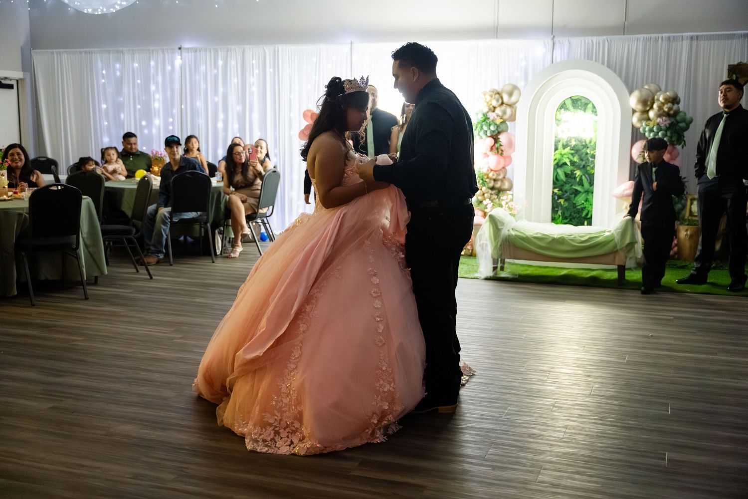 A young woman in a pink ball gown dances with a man in a suit at a quinceañera celebration.
