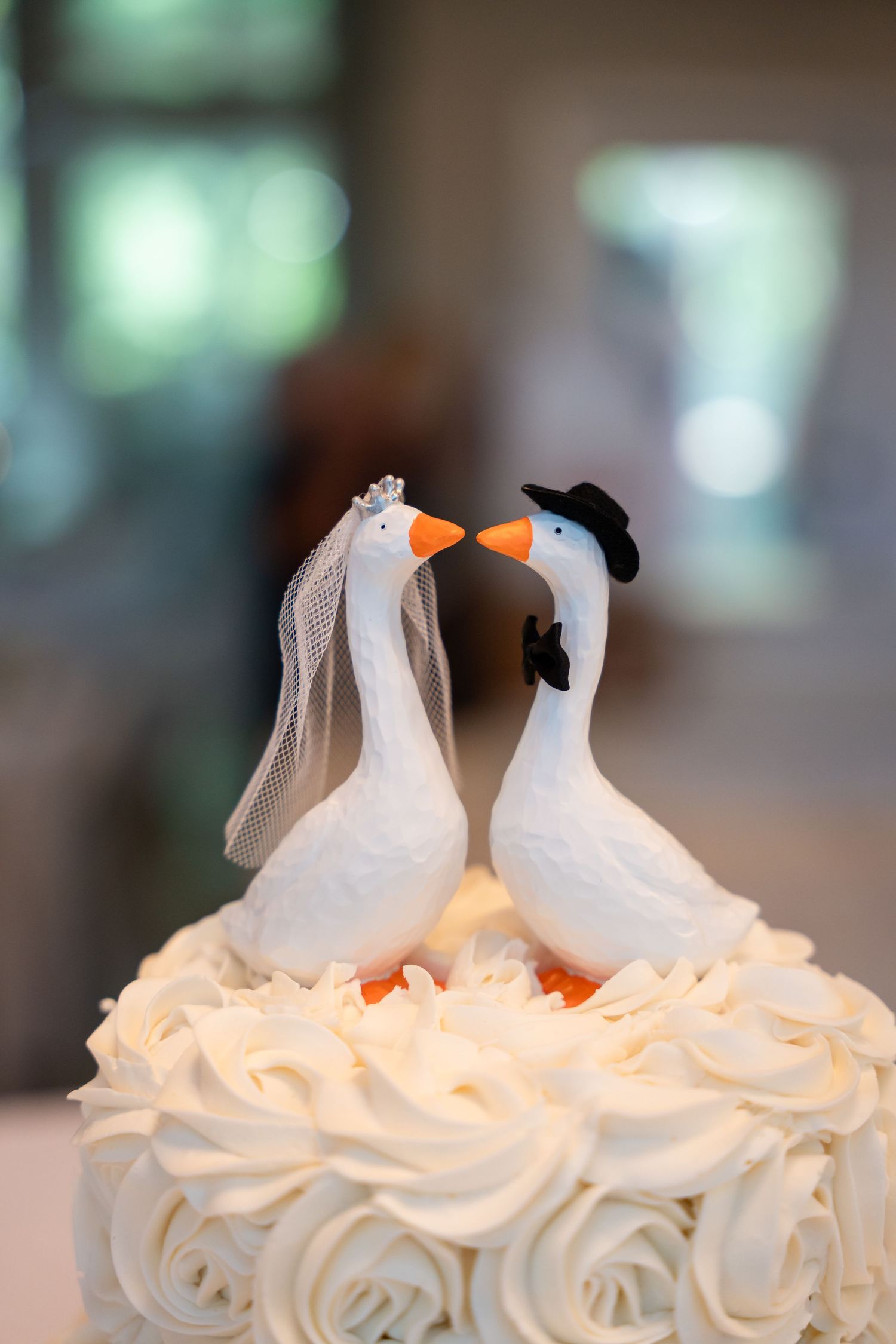 White wedding cake topped with two ceramic geese figurines dressed as bride and groom, kissing atop white rosette frosting.