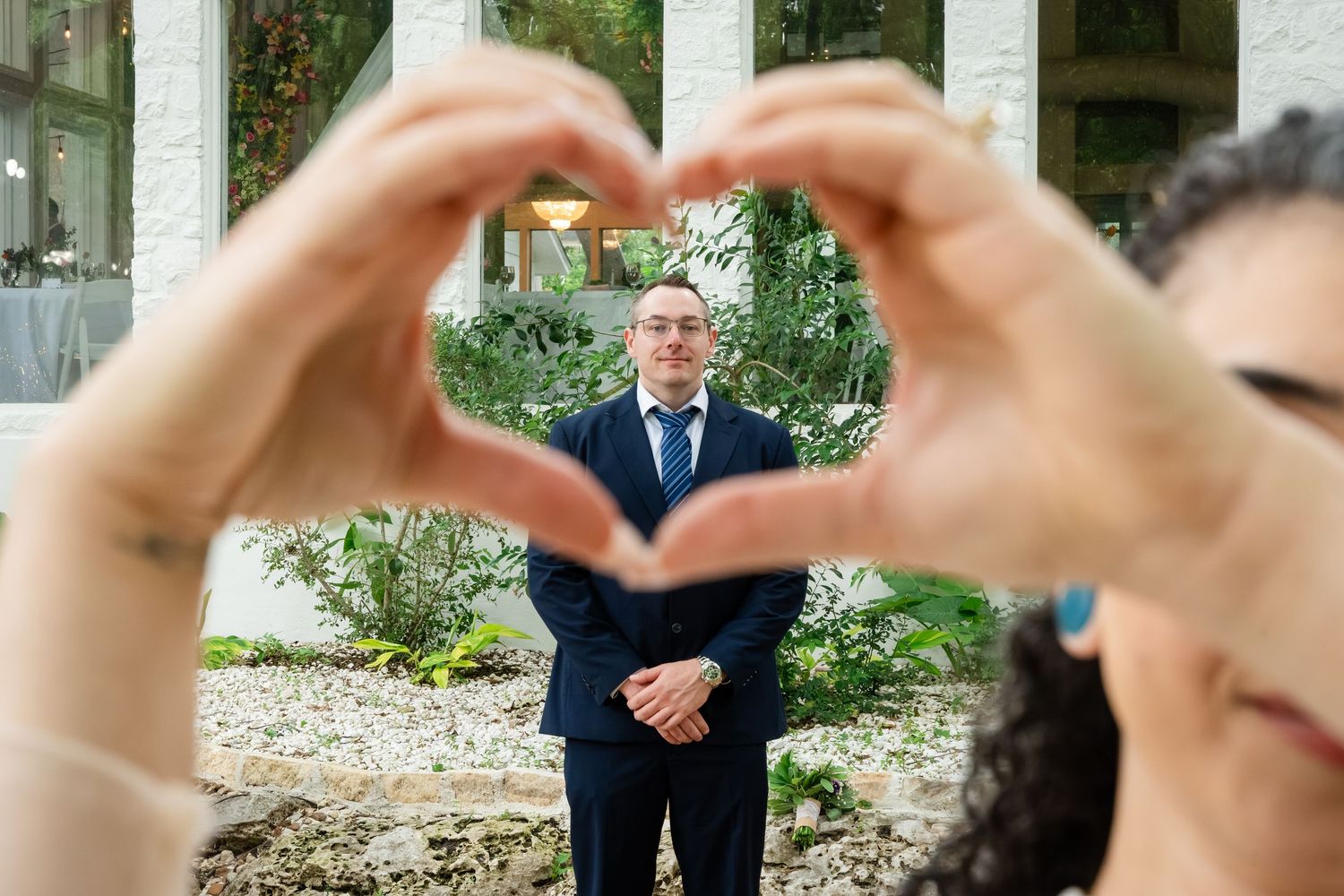 A groom in a navy suit is framed by two hands forming a heart shape at a wedding venue.