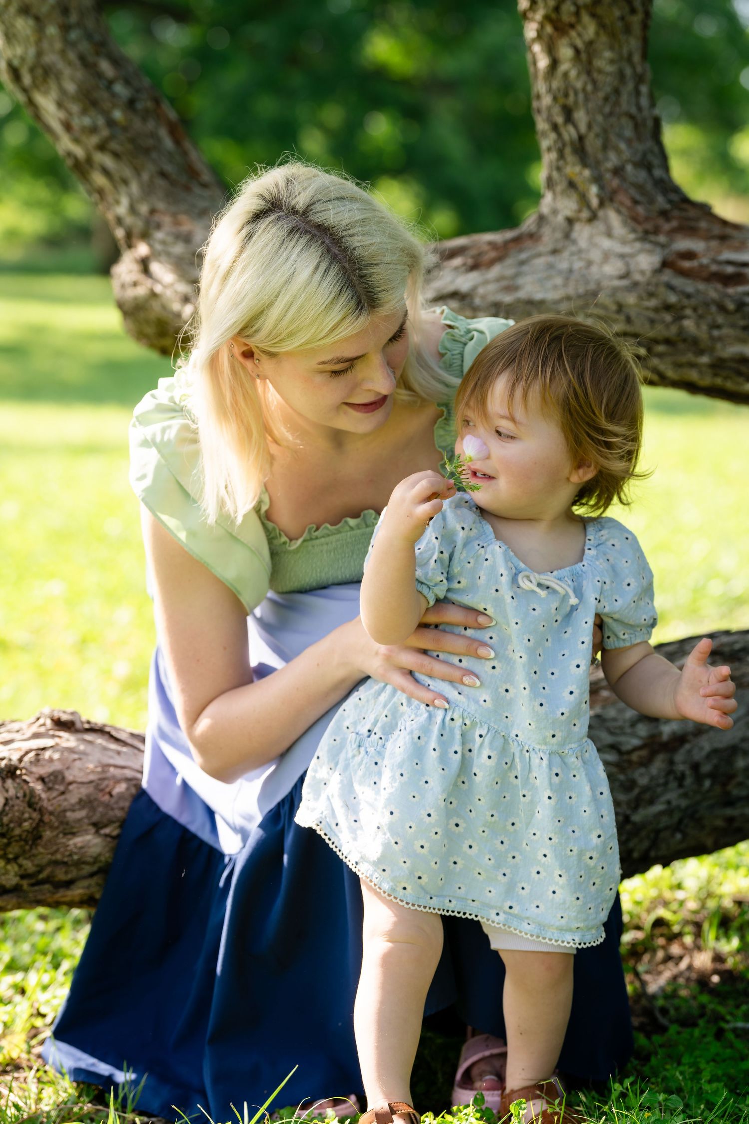 A blonde mother tenderly holds her toddler daughter in a sunny park, both wearing light summer dresses near a large tree.