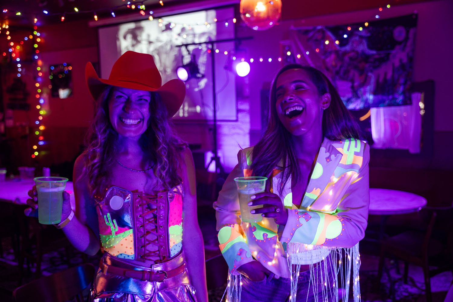 Two women in neon glowing outfits laugh at a blacklight party, one wearing a cowboy hat.
