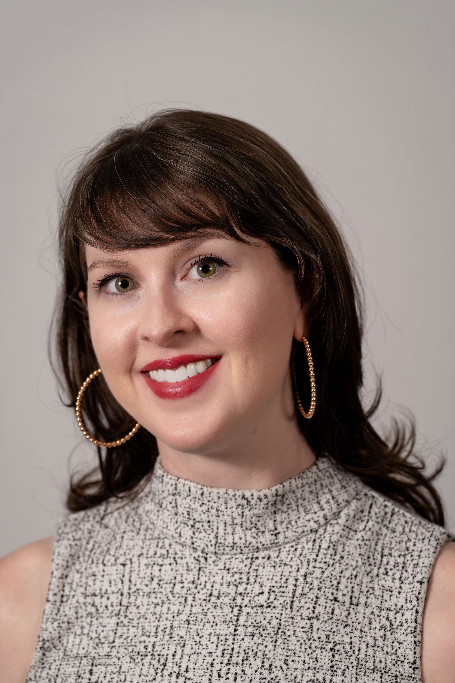 Smiling woman with dark hair, red lips, gold hoop earrings, wearing a gray sleeveless turtleneck top.
