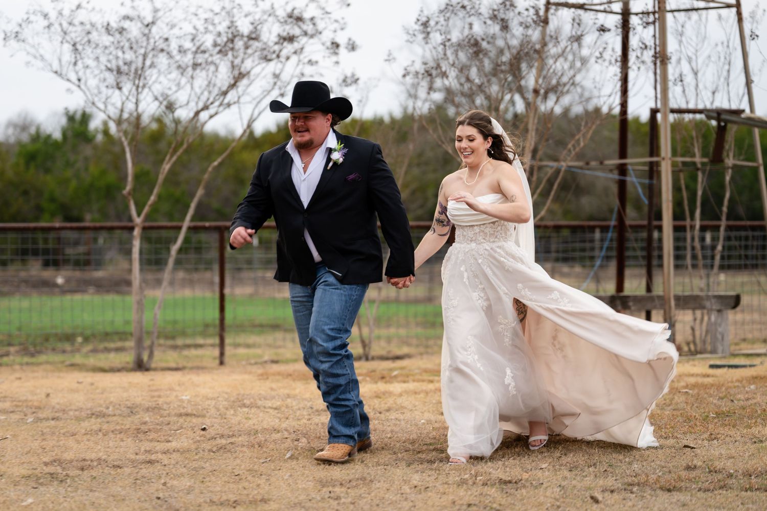 Bride in white gown and groom in black cowboy hat and jeans walk hand-in-hand at rustic outdoor ranch wedding.