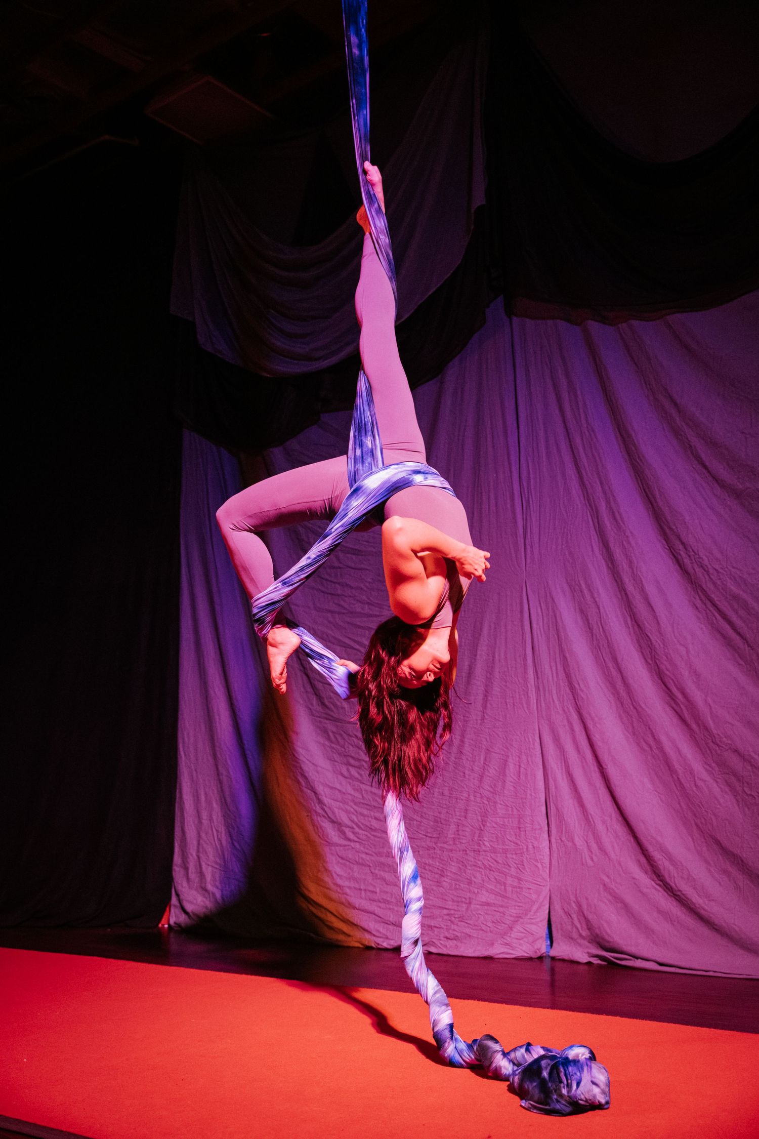 Aerial silk performer inverted on blue fabric on a red stage floor with dramatic purple lighting.