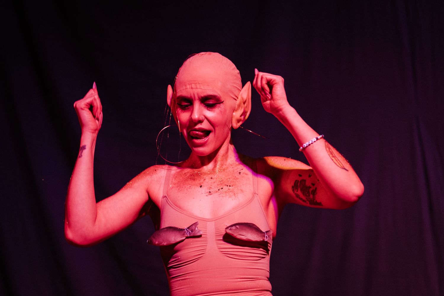 Bald performer in pink lighting wearing hoop earrings and a tan top, dancing on stage with tattoos visible.