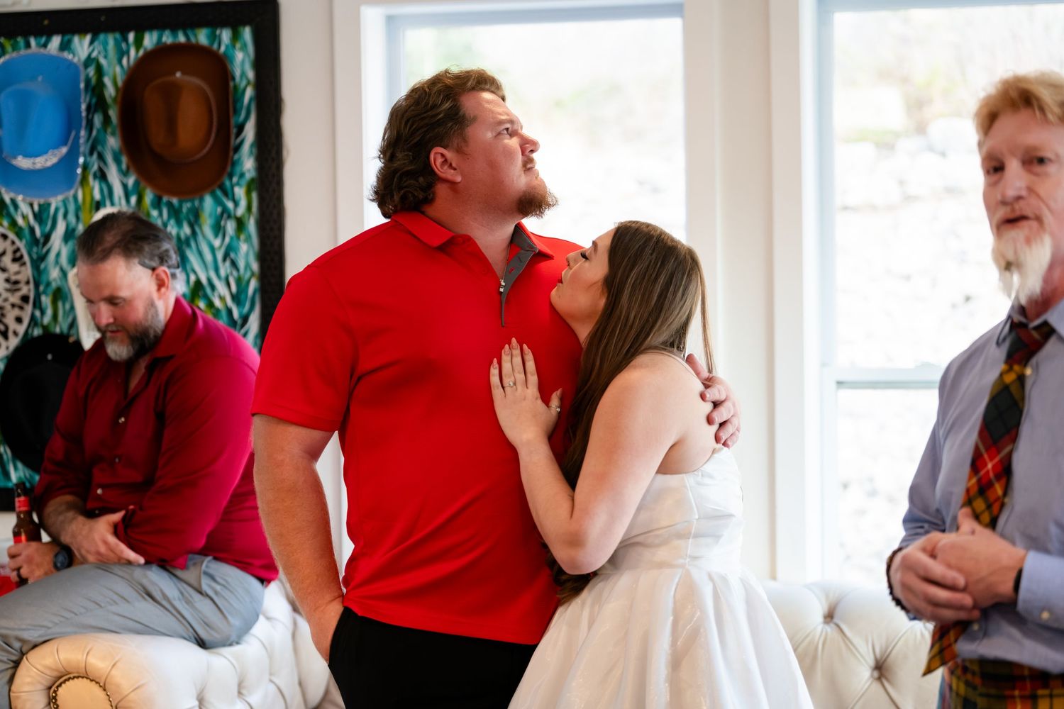 Bride in white dress kissing groom in red polo shirt at casual wedding celebration with guests in background.