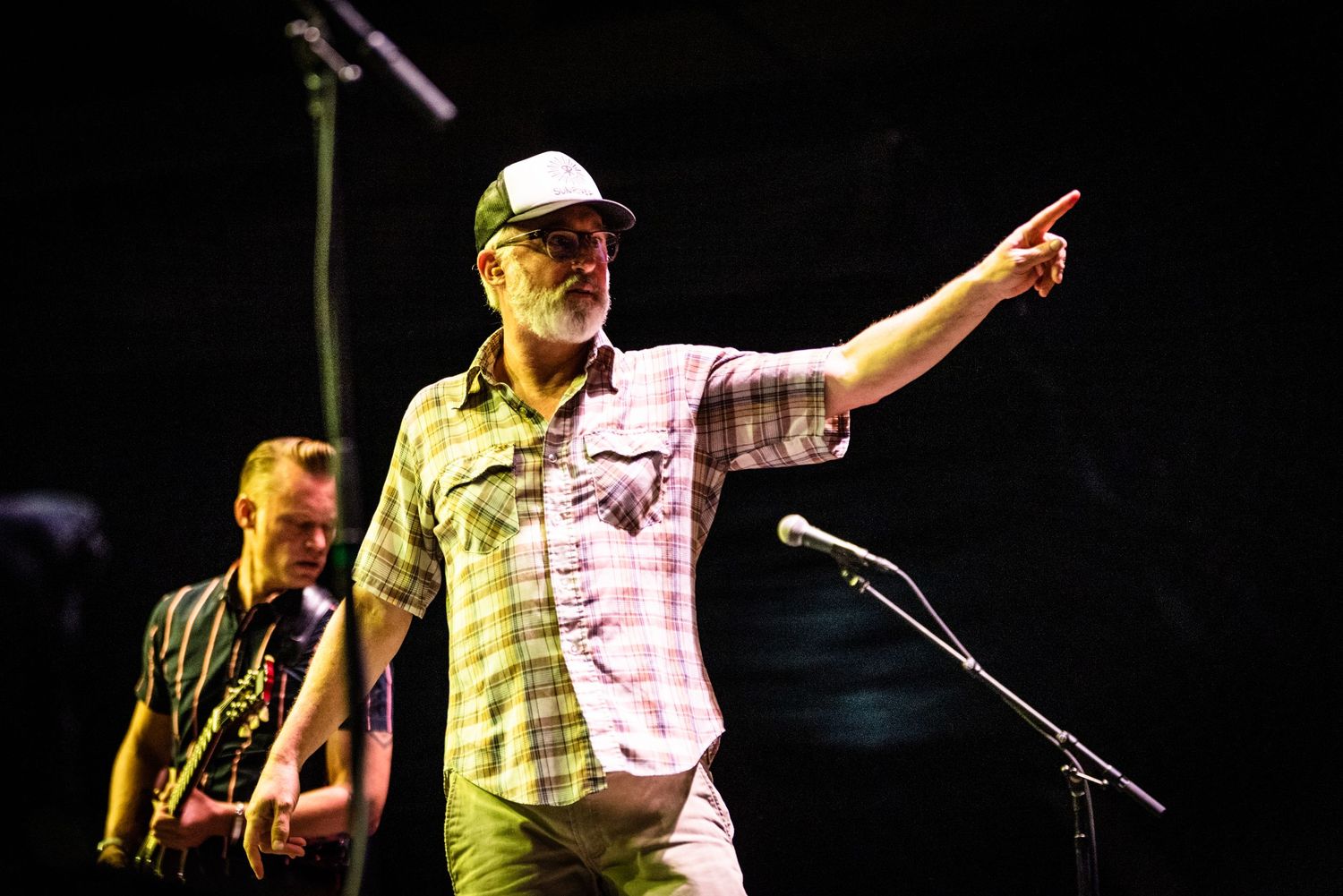 Performer in a white cap and plaid shirt gestures while singing on stage during a live musical performance.