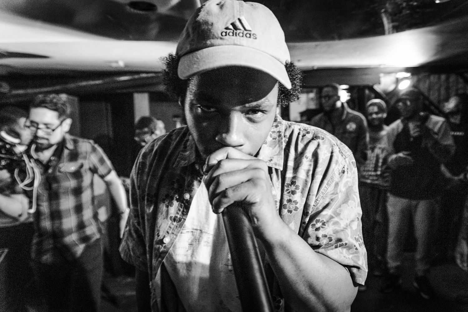 Black and white photo of a performer in an Adidas cap holding a microphone at a crowded live music event.
