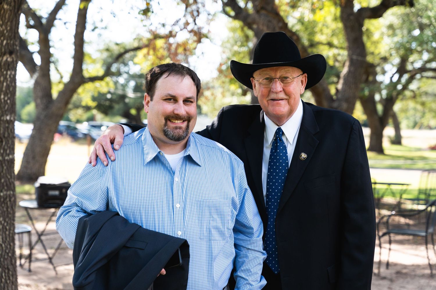 Two people in business attire pose together at an outdoor venue with chairs in background.