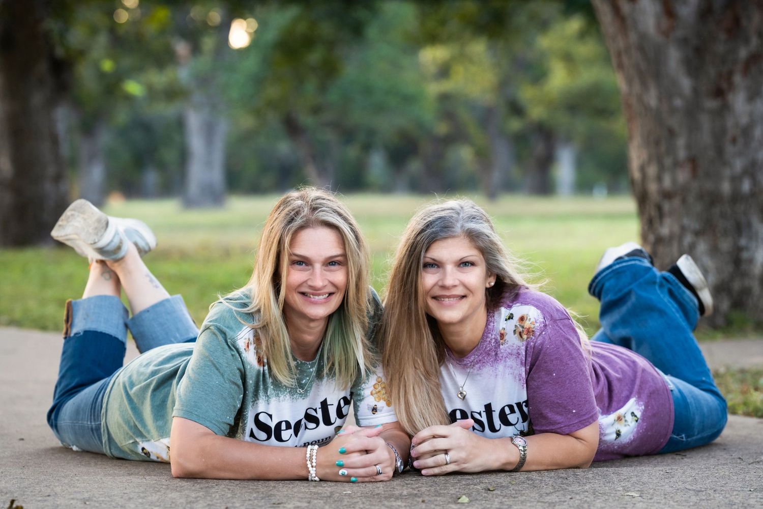 Two friends in matching graphic t-shirts lying on the ground in a park during a casual outdoor photoshoot.