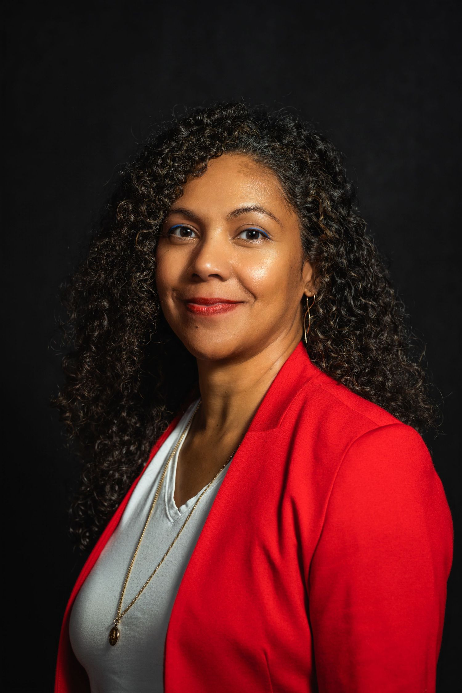 Studio headshot featuring person in bright red blazer with layered necklaces against black backdrop.