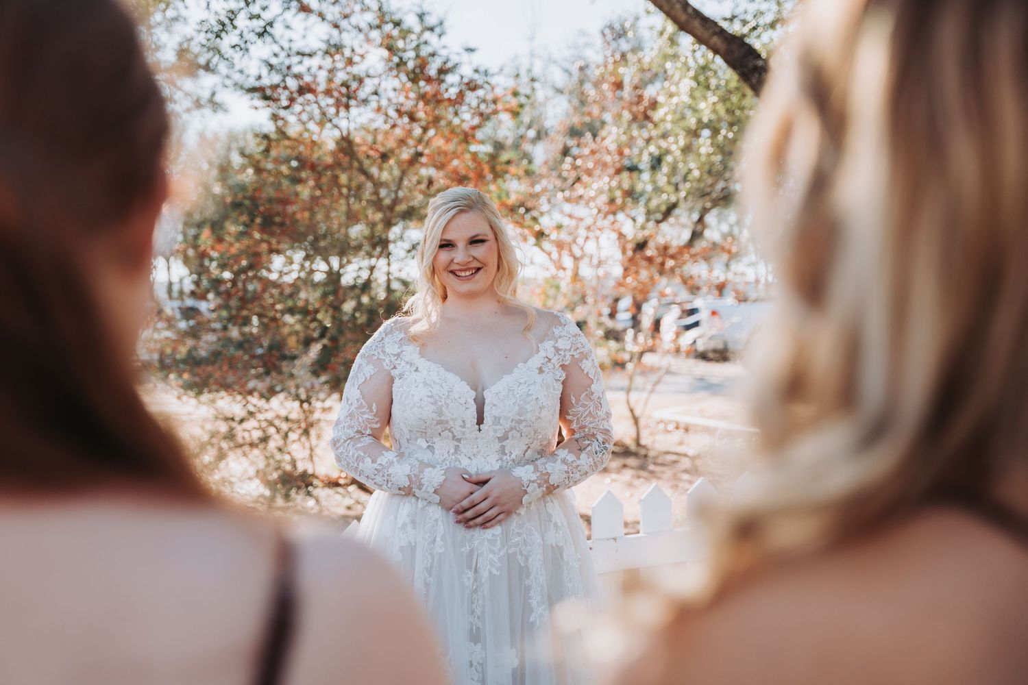A bride in a lace long-sleeve wedding gown stands outdoors during a sunny autumn ceremony.