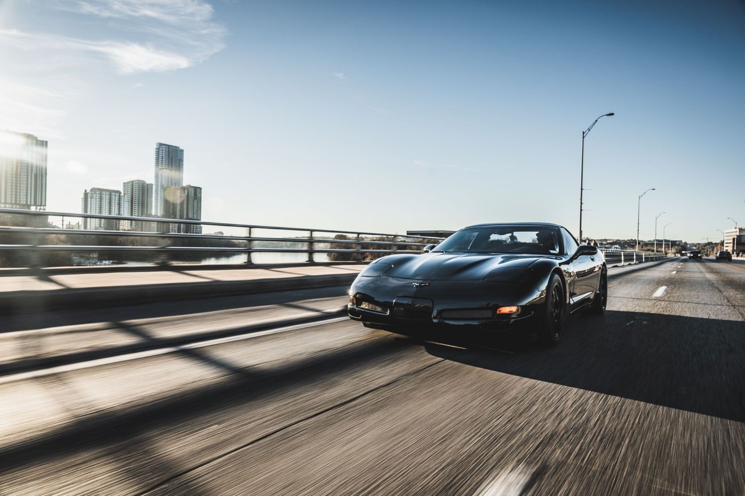 Black Chevrolet Corvette drives on elevated highway with city skyline visible in background.
