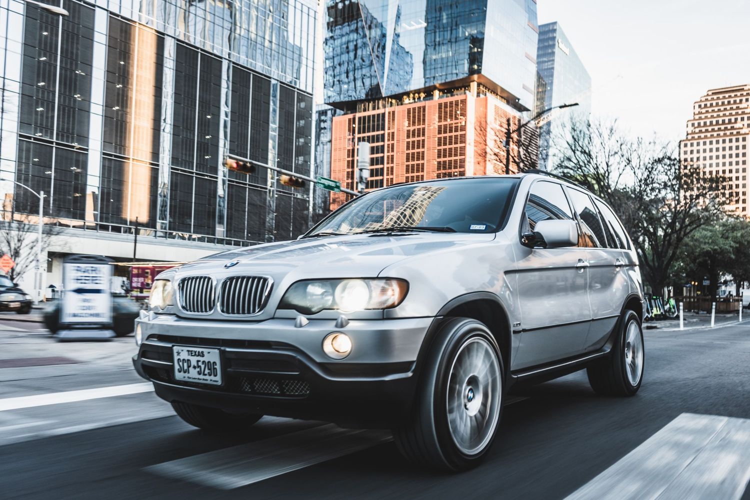 Silver BMW X5 SUV in motion on city street with modern office buildings in background.