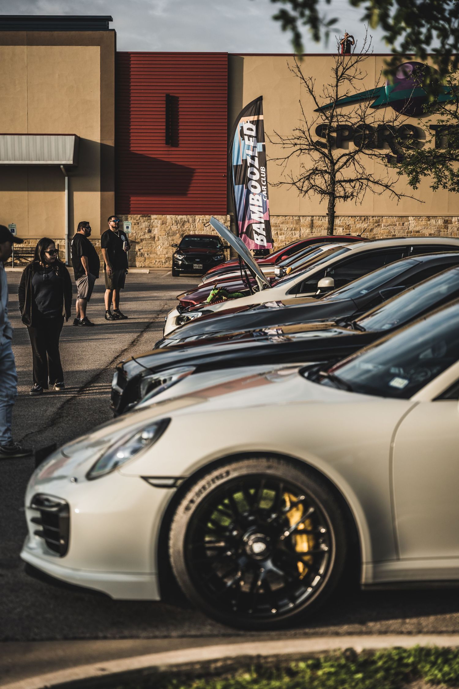 A line of luxury sports cars parked in front of a commercial building with a feather flag banner.
